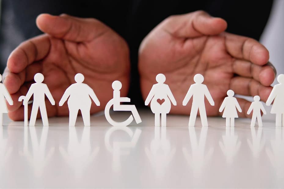 Sobre una mesa blanca hay unos muñecos muy minimalistas, solo figuras en blanco, de personas con bastón, gordas, con silla de ruedas, con un corazón en la barriga (embarazada), delgadas, niños, niñas, etc. De fondo las palmas de dos manos.