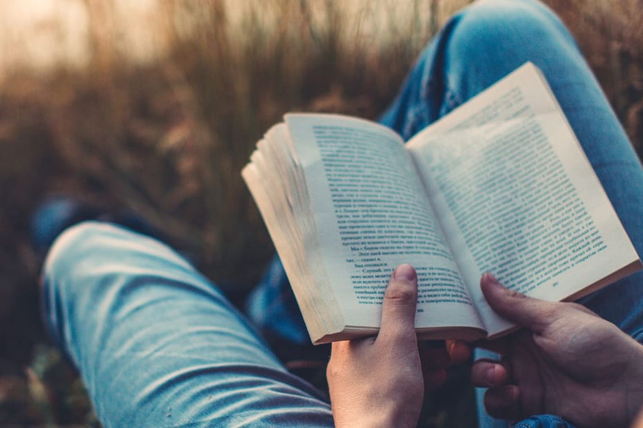 Fondo campo de espigas, se ven solo las piernas estiradas (lleva vaqueros) de alguien que sostiene entre sus manos un libro abierto y está leyendo. desde la esquina abajo a la derecha hacia la contraria.